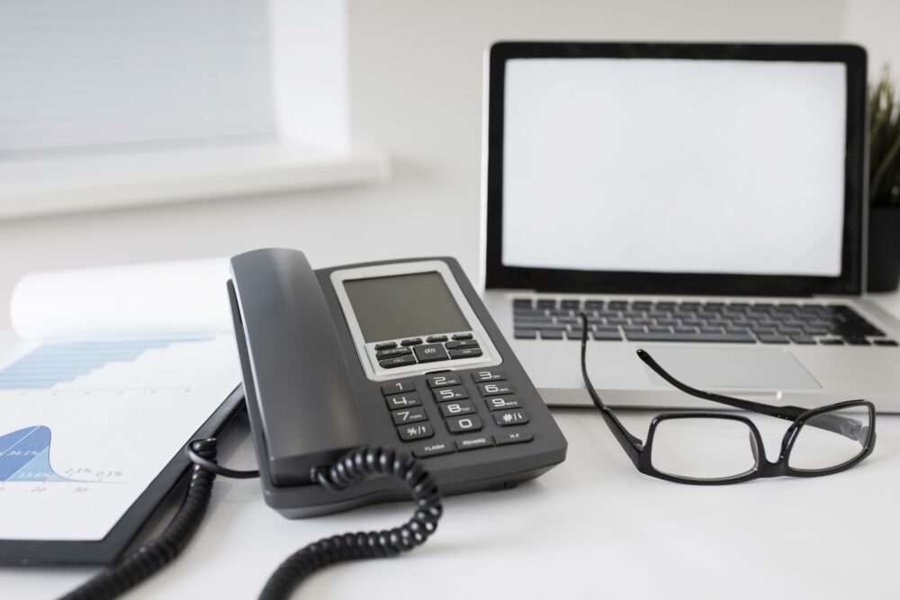 A laptop, eyeglasses, and a document with graphs are neatly arranged on the desk alongside a corded device characteristic of on-premise phone systems.