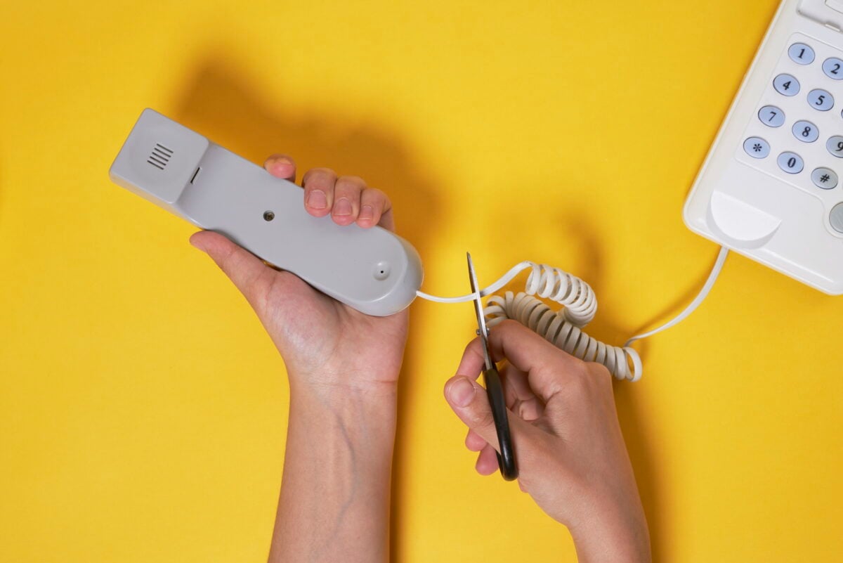 Amidst a vibrant yellow backdrop, a person decisively cuts the cord of a landline telephone with scissors, symbolizing the era of ISDN Switch Off.